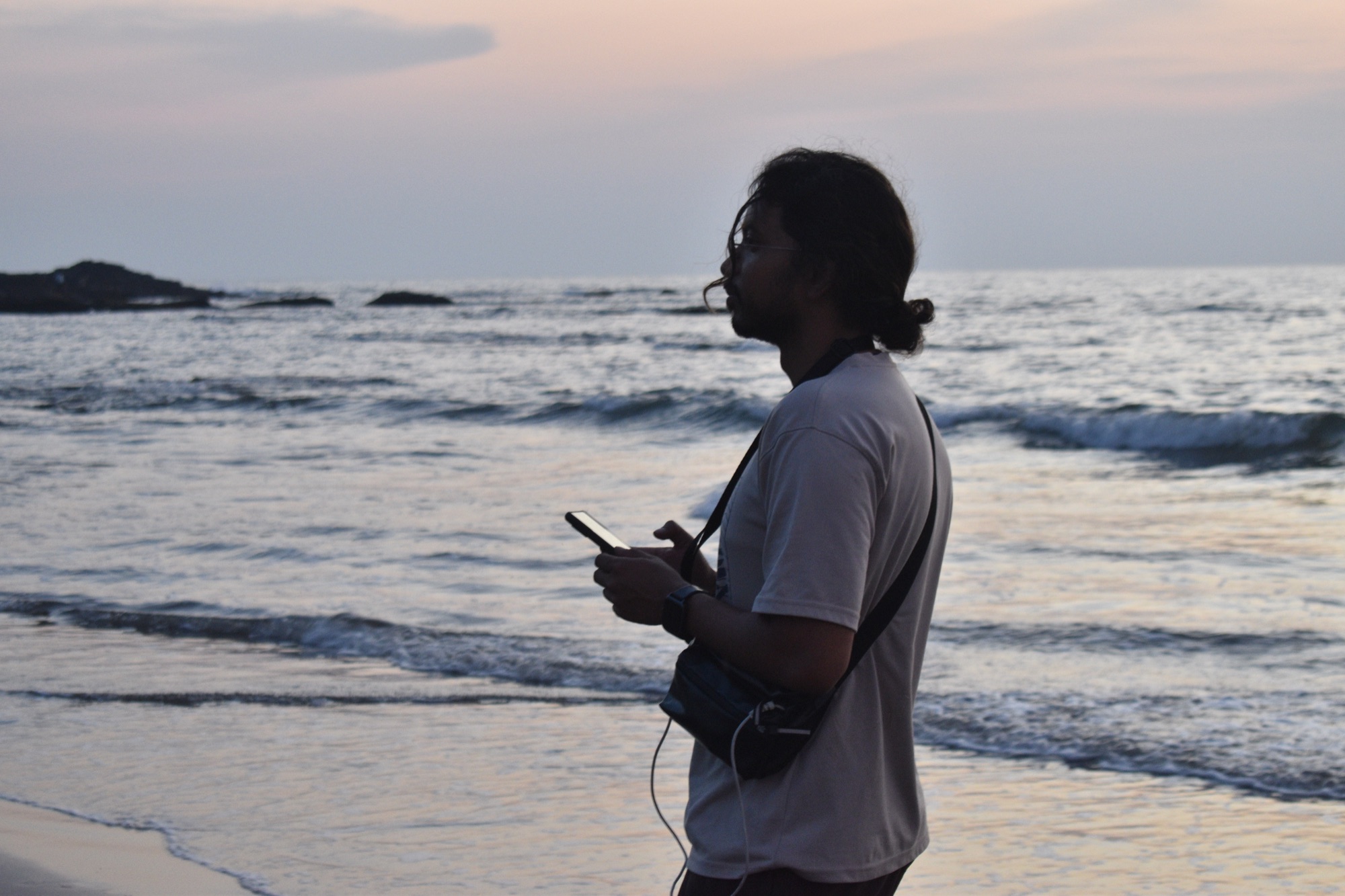 Aayush on a beach at golden hour, camera bag slung across, sea softly breaking behind